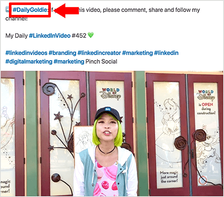 Αυτό είναι ένα στιγμιότυπο οθόνης που δείχνει πώς η Goldie Chan χρησιμοποιεί hashtag στο κείμενο των αναρτήσεων βίντεο στο LinkedIn. Οι κόκκινες επεξηγήσεις δείχνουν το hashtag #DailyGoldie στο κείμενο, το οποίο είναι μοναδικό στις αναρτήσεις βίντεο και τη βοηθά να παρακολουθεί τις κοινοποιήσεις. Η ανάρτηση περιλαμβάνει επίσης άλλα σχετικά hashtag που βοηθούν τους χρήστες να βρουν το βίντεό της, συμπεριλαμβανομένου του #LinkedInVideo. Στην εικόνα βίντεο, η Goldie στέκεται μπροστά σε μερικές πόρτες σε μια οθόνη του κόσμου της Disney. Είναι ασιατική γυναίκα με πράσινα μαλλιά. Φορά ένα μαύρο καπάκι LinkedIn, ένα μαύρο κολιέ choker, ένα ροζ πουκάμισο με μακαρόν και ένα μπλε και άσπρο σακάκι.